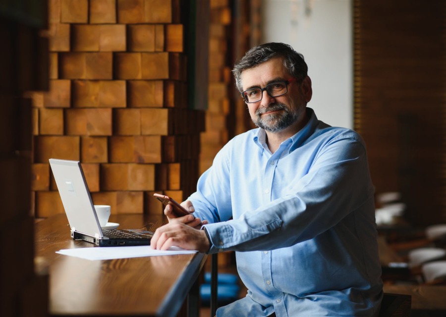 Homem de óculos usando celular e notebook em um café