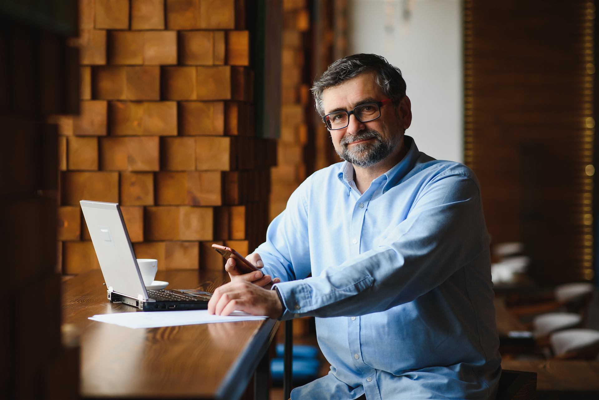 Homem de óculos usando celular e notebook em um café